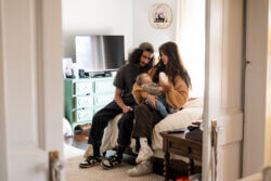 A young couple sitting on a bed together bottle-feeding their baby.