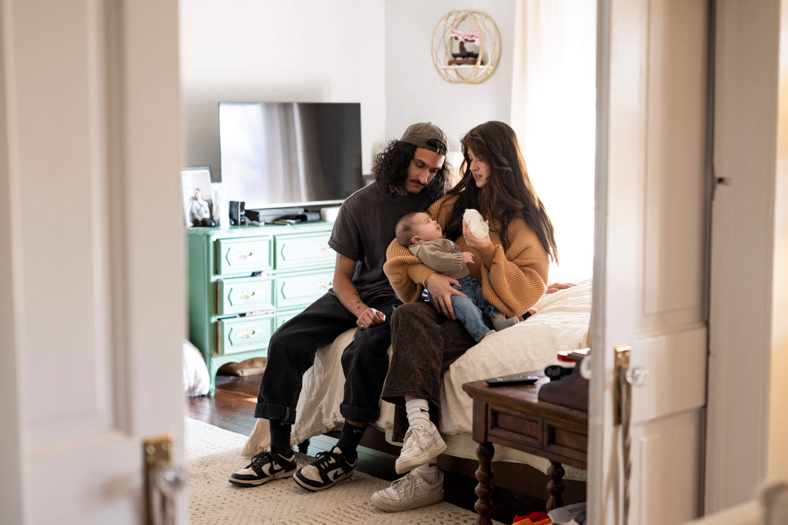 A young couple sitting on a bed together bottle-feeding their baby.