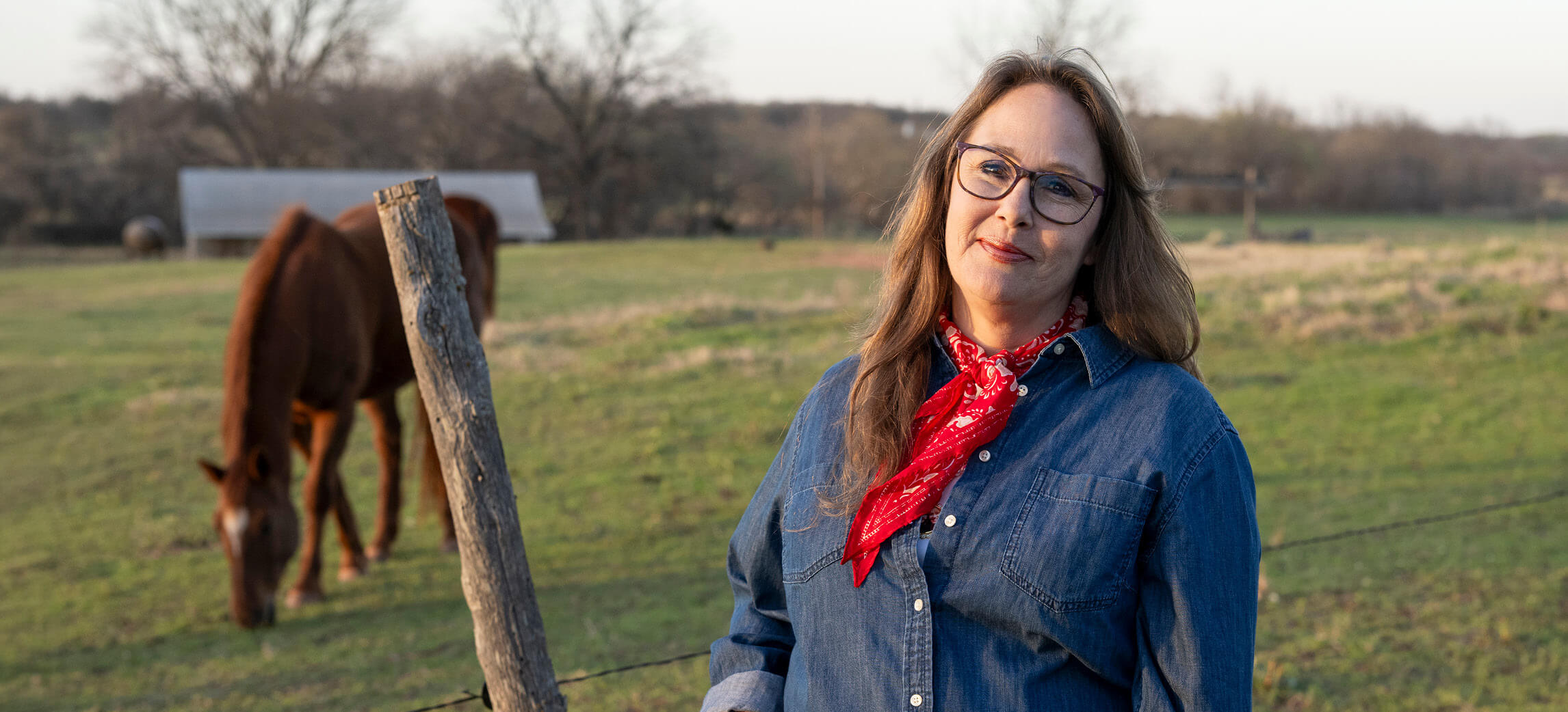 A woman wearing glasses and a red neckerchief, reminiscent of an Oklahoma Tobacco Helpline representative, stands in a pasture near a grazing horse.